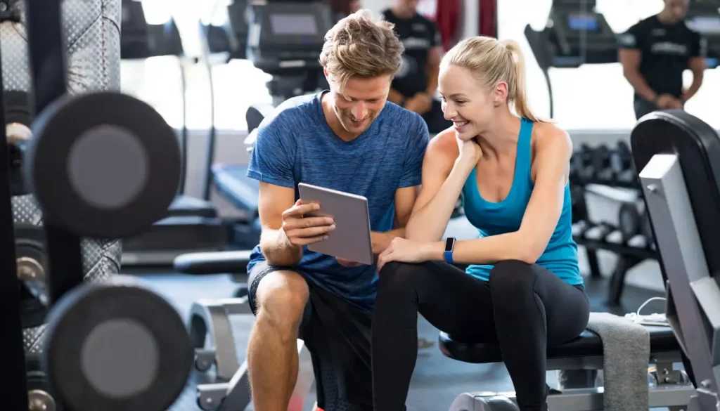 Effektiv abnehmen mit App – Dein Weg zur Fitness! 3 Frau und Mann im Fitnessstudio betrachten gemeinsam die Yukado App auf einem Tablet während des Workouts